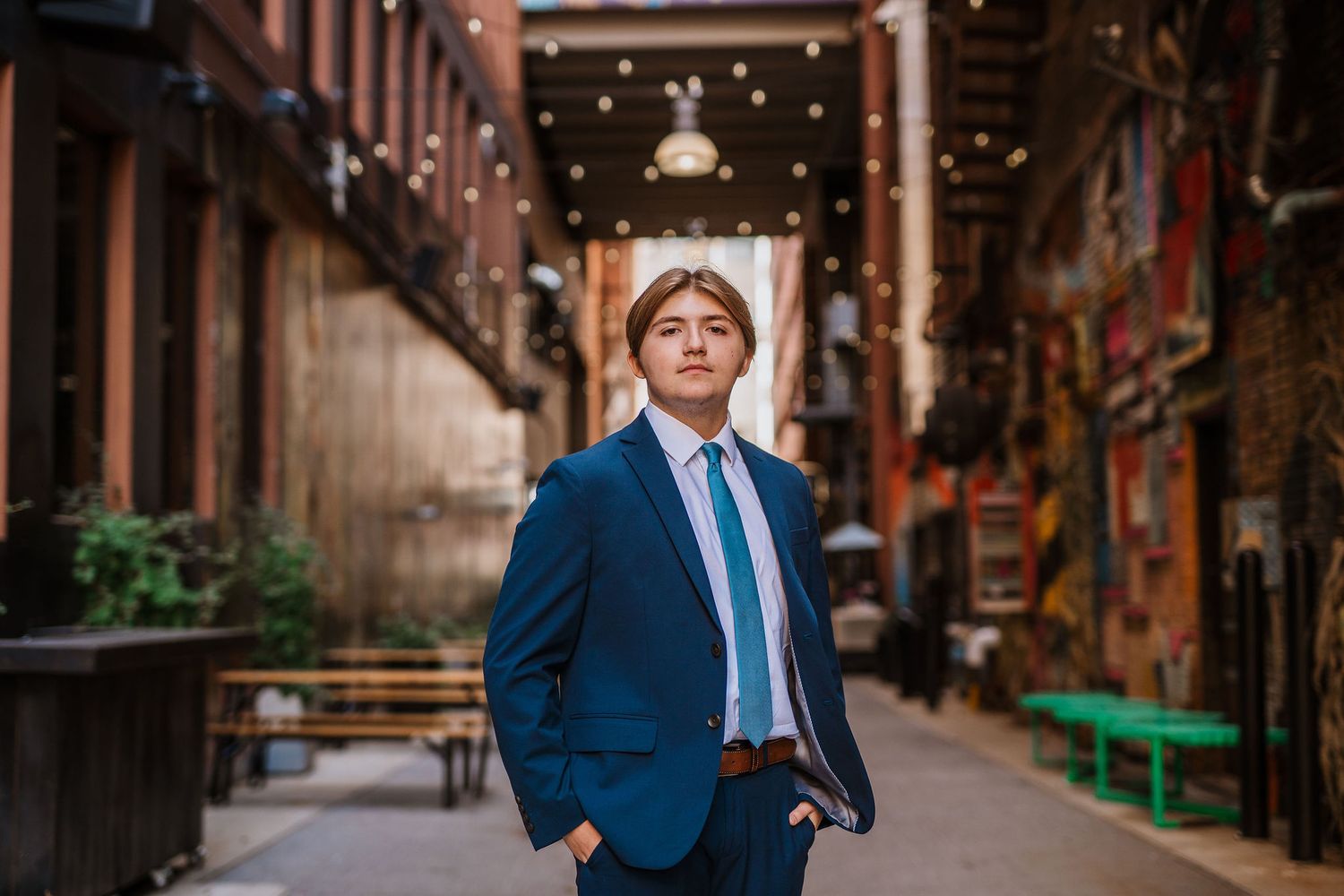 Young man in blue suit and tie posing confidently in an urban brick alley with string lights.