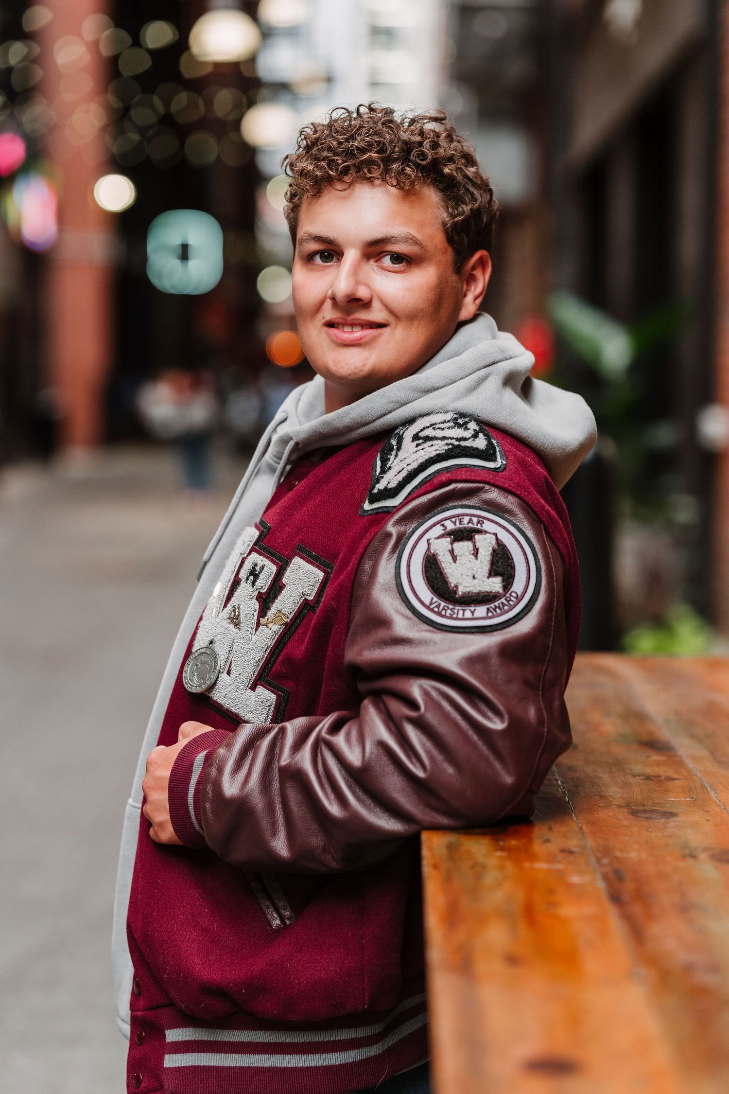 Young person in a maroon varsity jacket with patches, leaning on a wooden table in an urban alleyway.