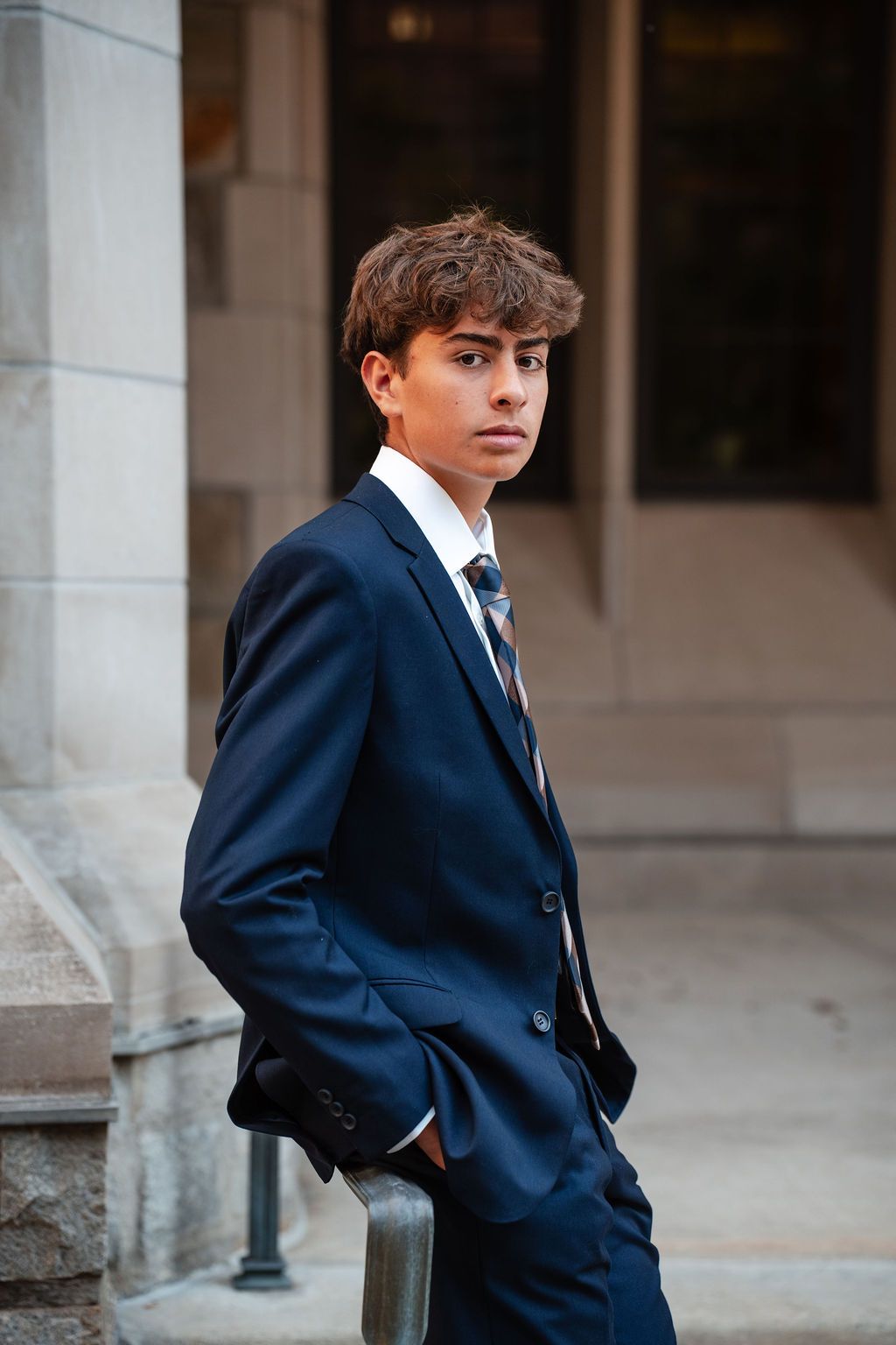 Young man in navy suit and tie posing confidently outdoors near stone columns.