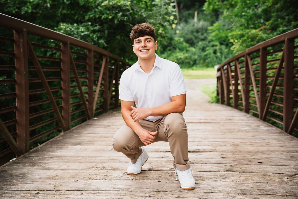 Young man in white polo and khaki pants crouching on a wooden bridge surrounded by lush green foliage.