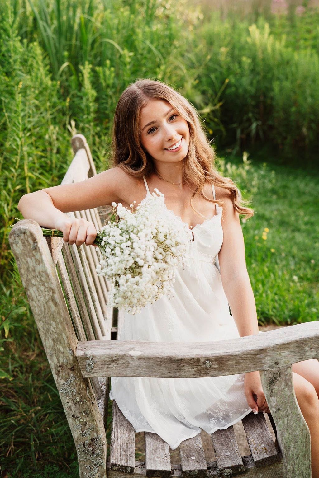 A dreamy outdoor portrait featuring a white sundress and wildflowers on a rustic wooden bench in a garden setting.