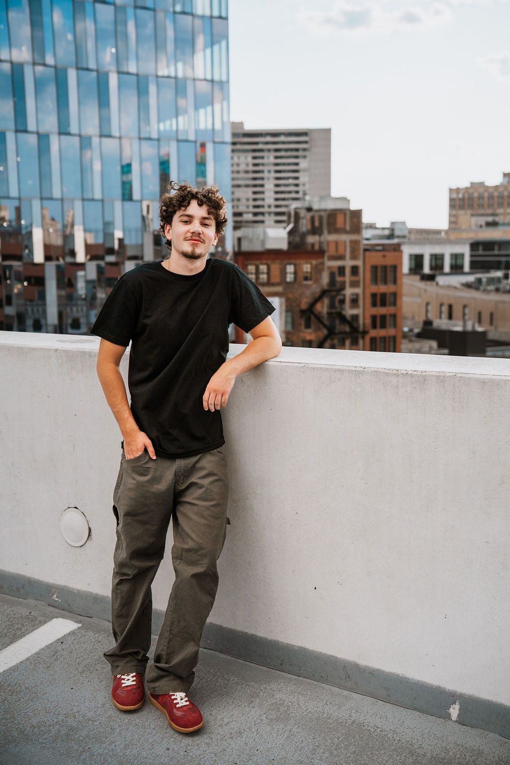 A person in a black t-shirt and gray pants poses on an urban rooftop with city buildings in the background.