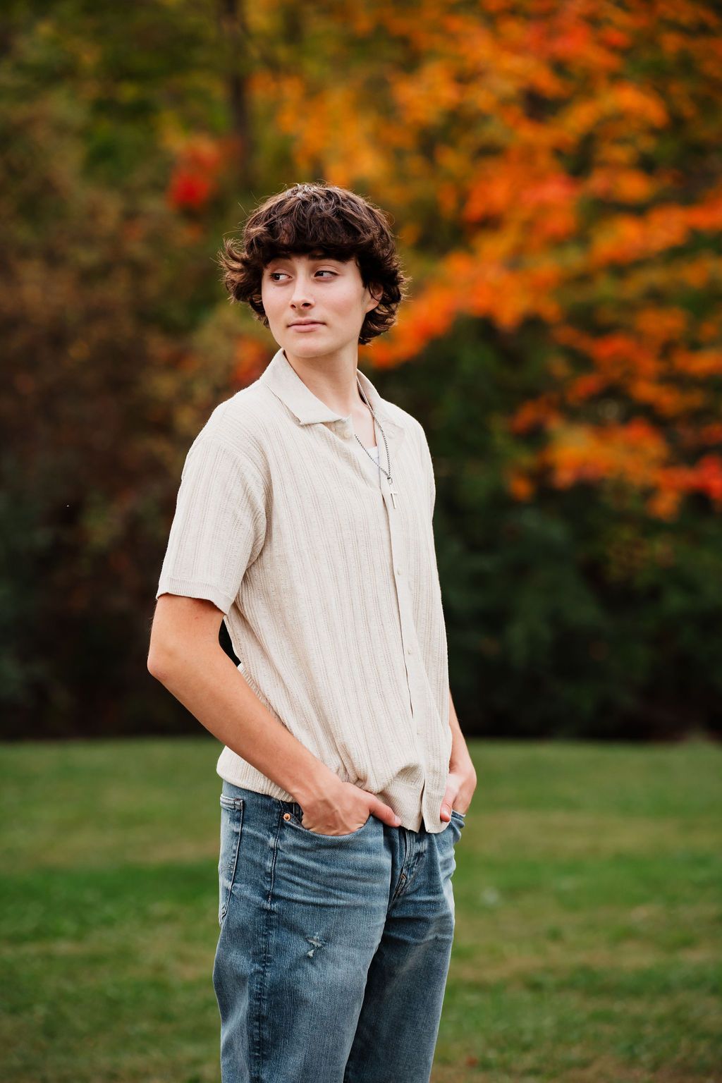 Teen boy in beige shirt and jeans poses outdoors with vibrant autumn foliage in background.