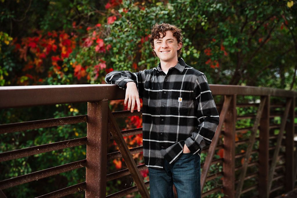 Young man in plaid shirt leaning on wooden bridge railing with colorful autumn foliage backdrop.
