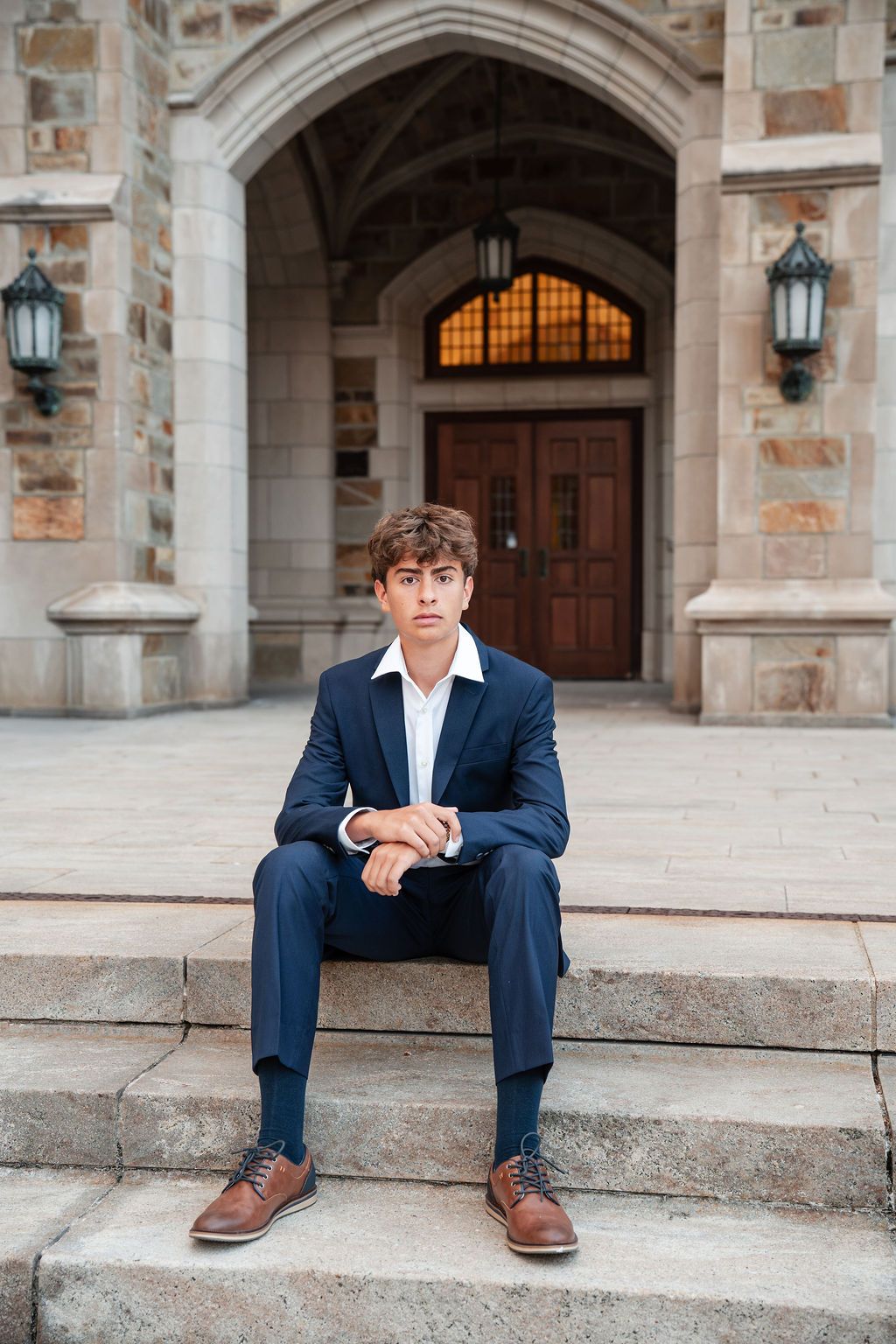 Young man in navy suit and brown dress shoes sitting on stone steps outside an elegant building.