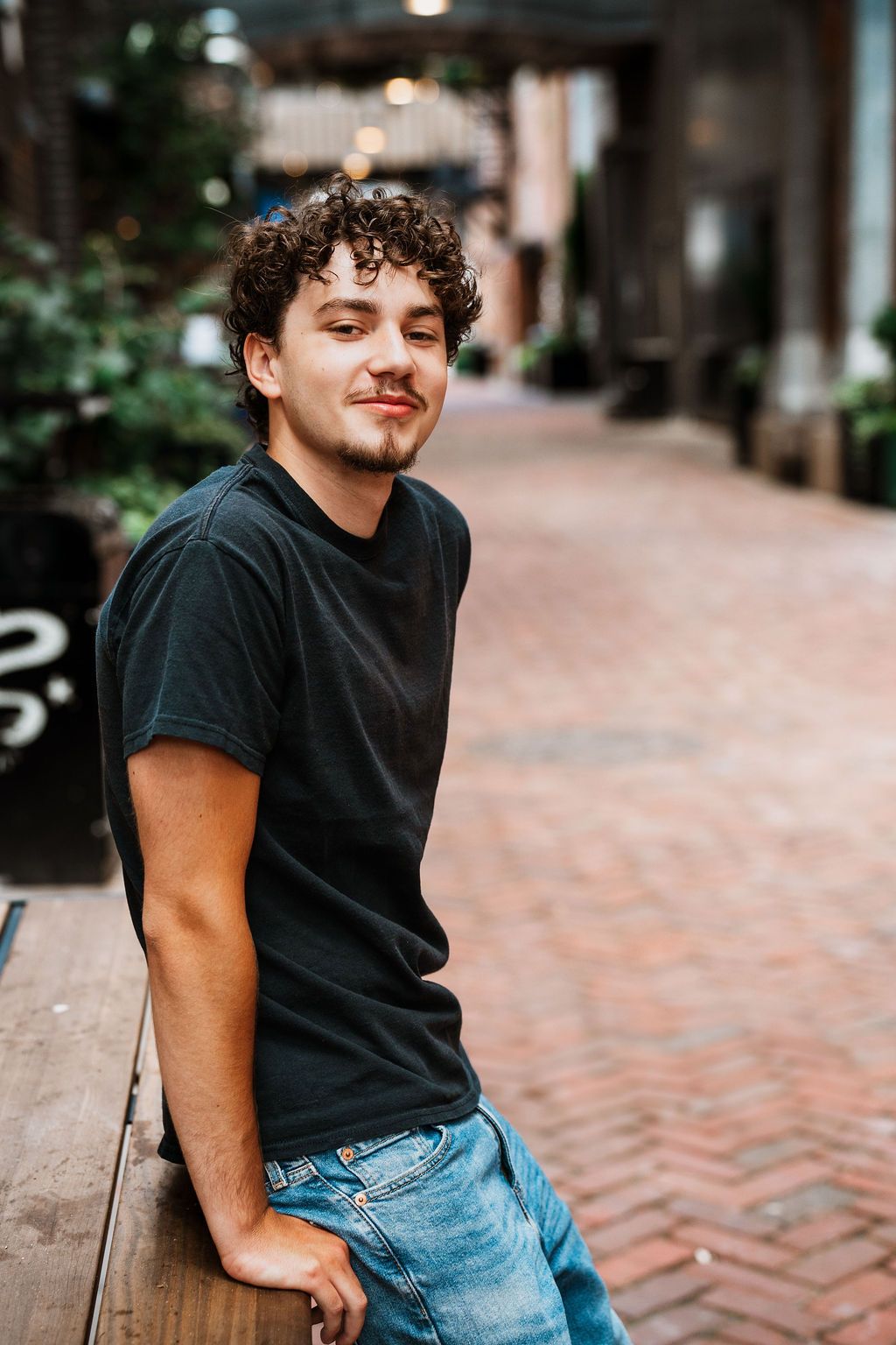 Young man with curly hair in black t-shirt and jeans leaning on bench in brick alley.