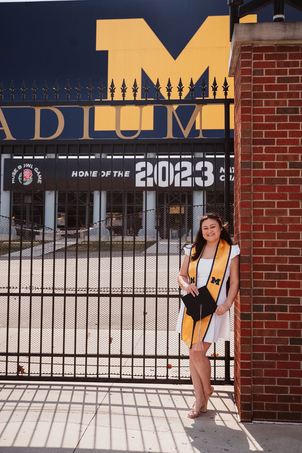 University of Michigan 2023 graduate posing by stadium gate in white dress with maize stole.