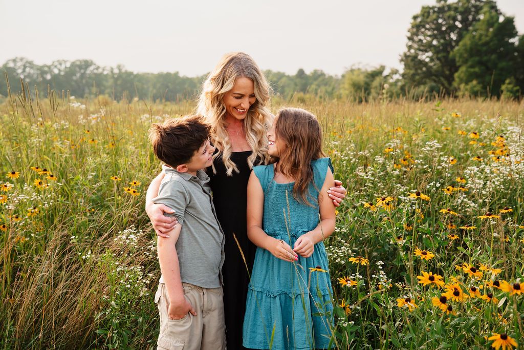 A mother smiles with her two children in a sunny wildflower field filled with yellow black-eyed Susans.