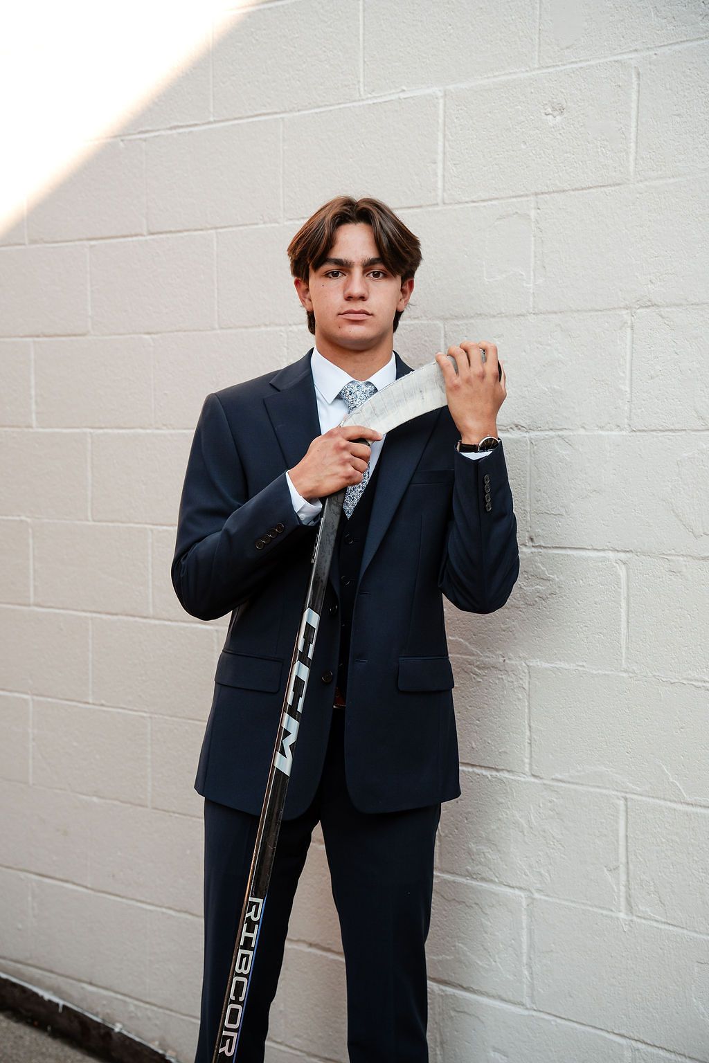Student poses stylishly in dark suit and tie against white wall background.