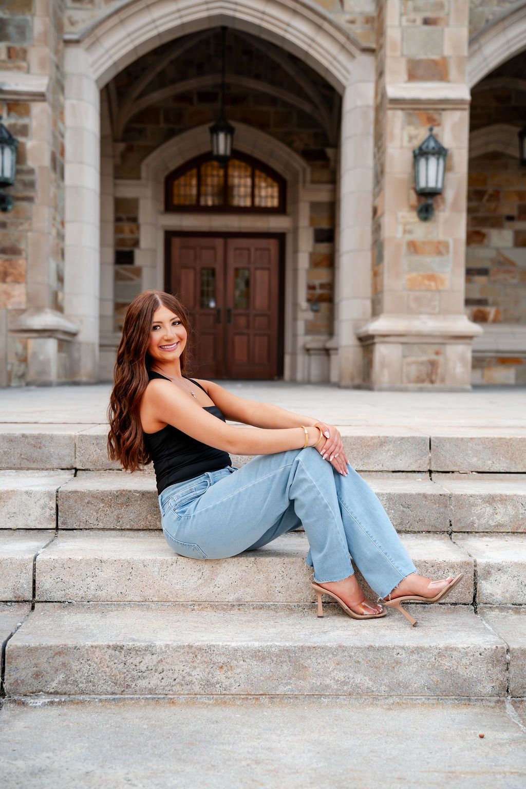 A person in a black top and light jeans sits casually on stone steps in front of an ornate archway entrance.