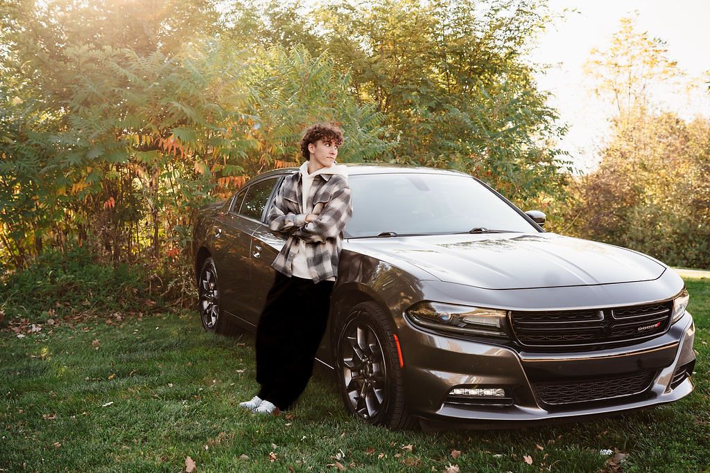 Teen boy in plaid shirt leaning against a dark gray Dodge Charger surrounded by fall foliage.