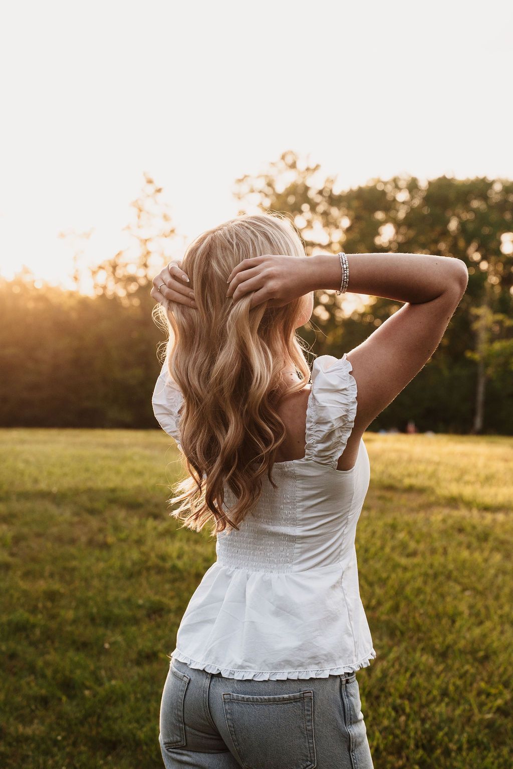 Blonde woman in white top and jeans stands in sunlit field at golden hour, hands in hair, back to camera.