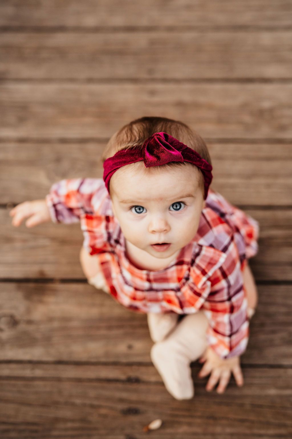 Blue-eyed baby girl in plaid dress and red bow headband sitting on wooden floor looking up.