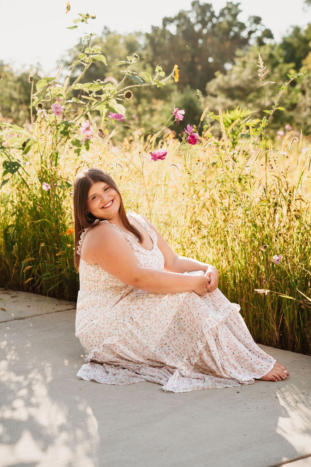 Young woman in floral maxi dress sits smiling on path surrounded by wildflowers in golden sunlight.
