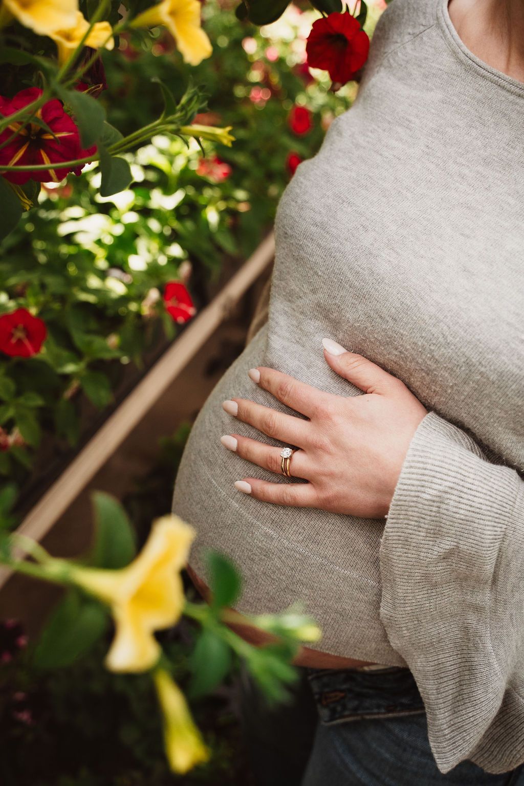 Pregnant woman in gray sweater gently holding her baby bump, surrounded by colorful red and yellow flowers.
