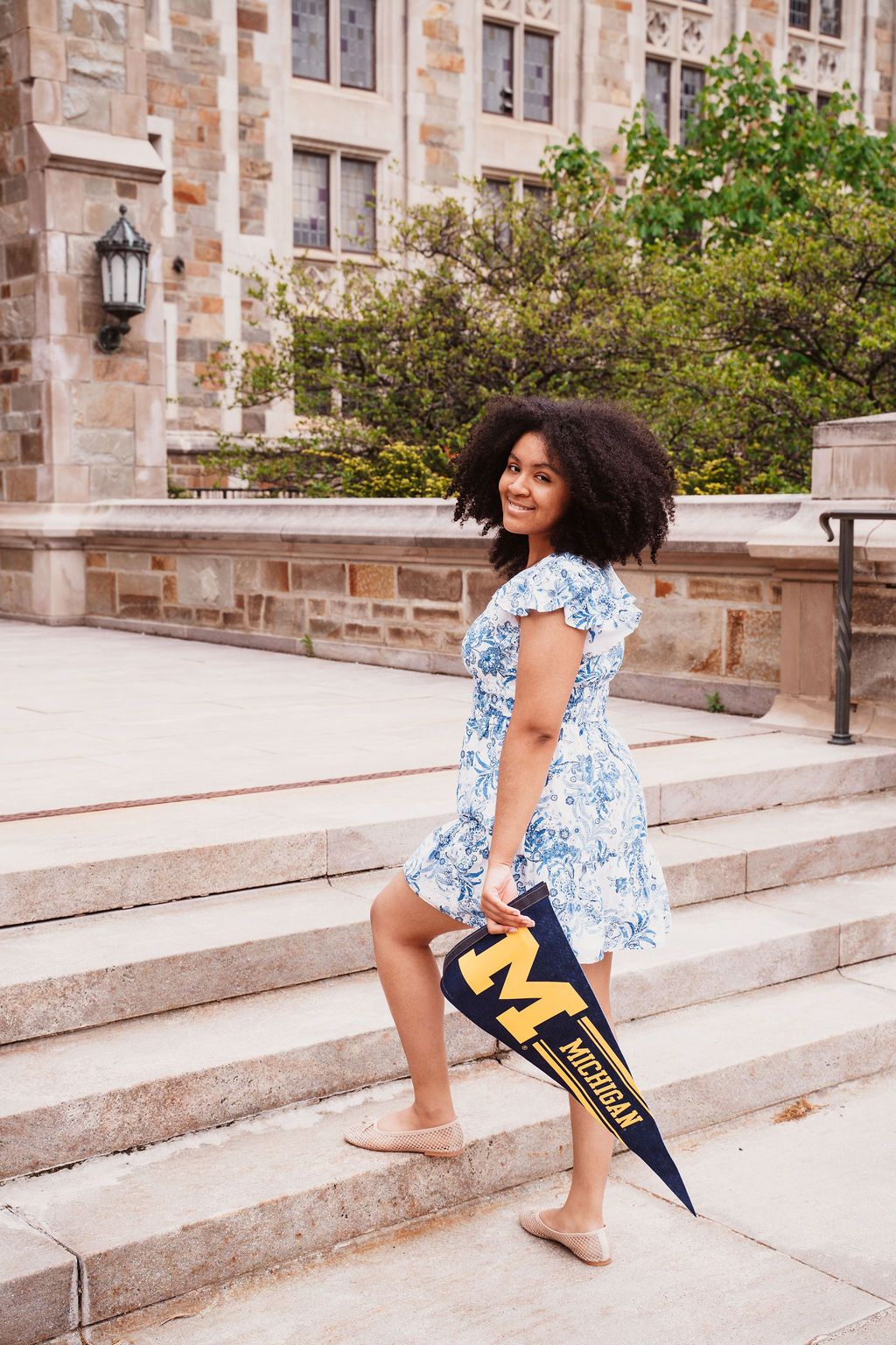 Young woman in floral dress holding University of Michigan pennant on stone steps of campus building.