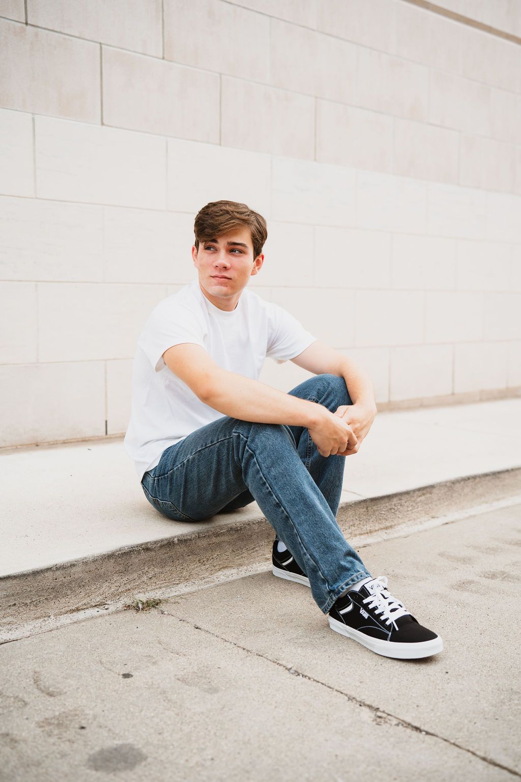 Young man in white t-shirt and jeans sitting casually against a light stone wall outdoors.
