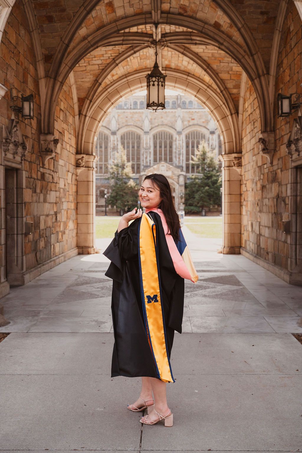 Graduate in black gown with Michigan maize stole poses under stone arches at University of Michigan.
