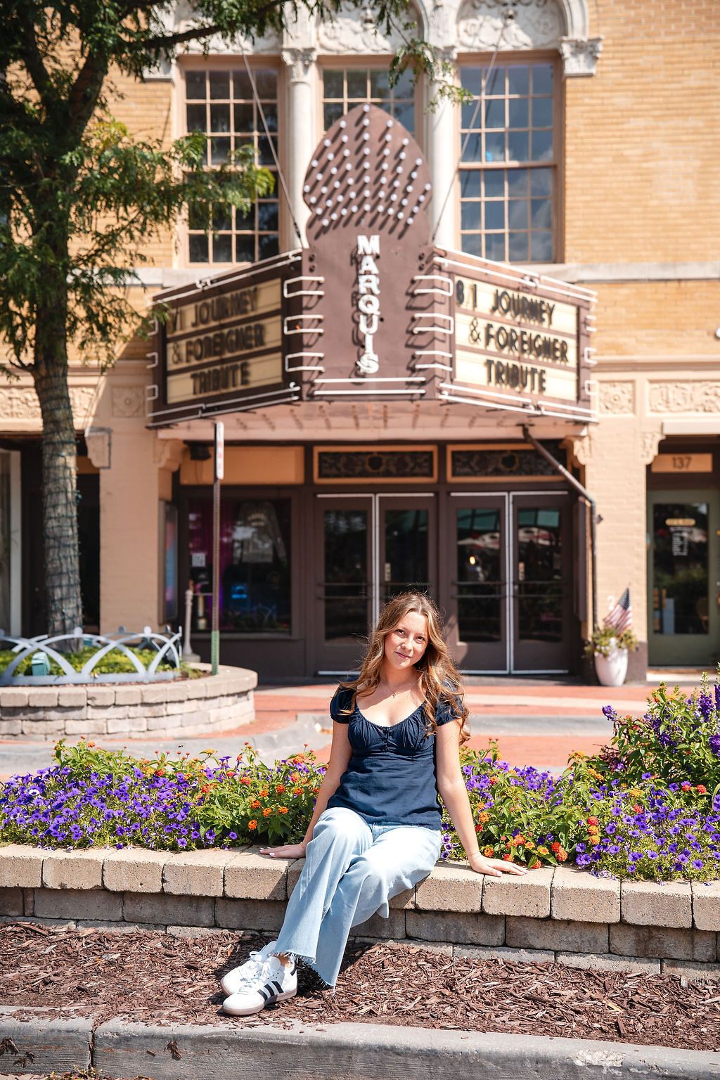 Young woman sitting on a flower bed in front of the Marqui Theater advertising a Journey and Foreigner Tribute show.