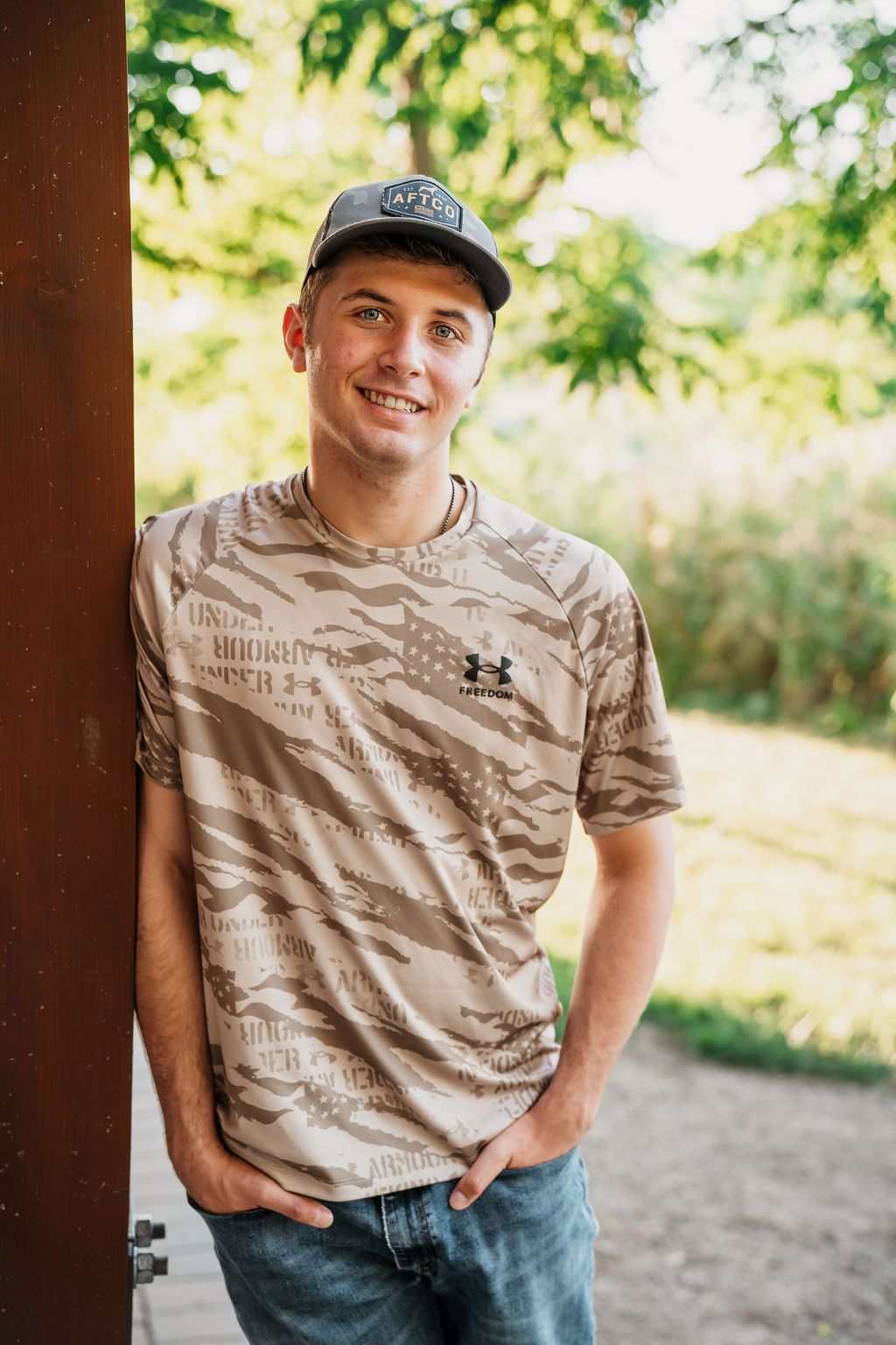 Young man smiling in camo Under Armour shirt and cap, leaning against a wooden post outdoors.