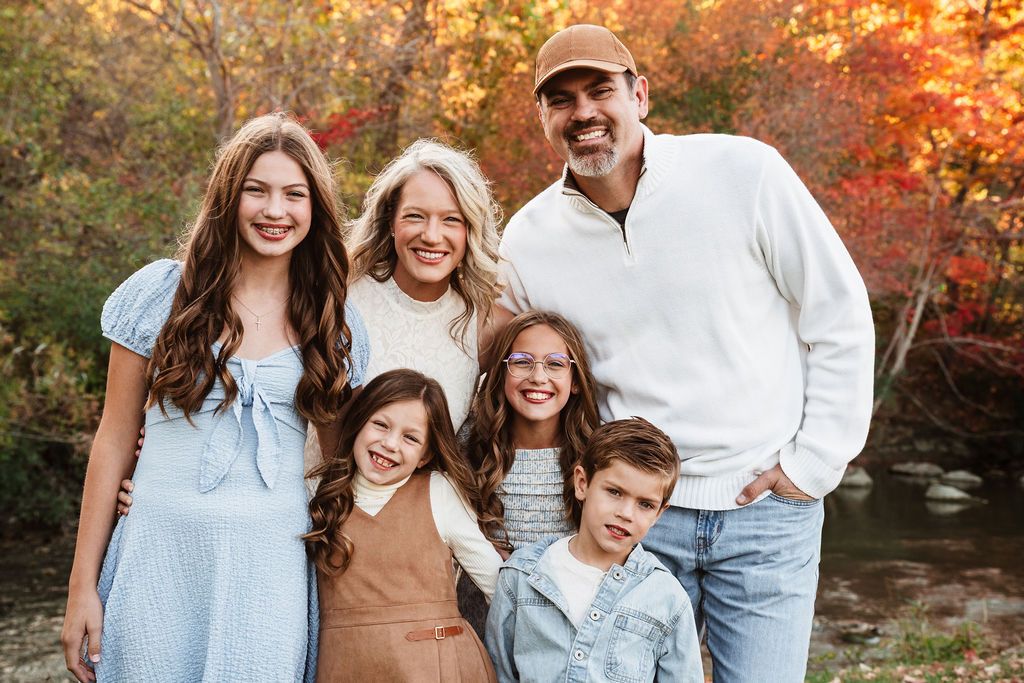 A smiling family of six poses outdoors among vibrant fall foliage, featuring parents and four children in coordinated outfits.