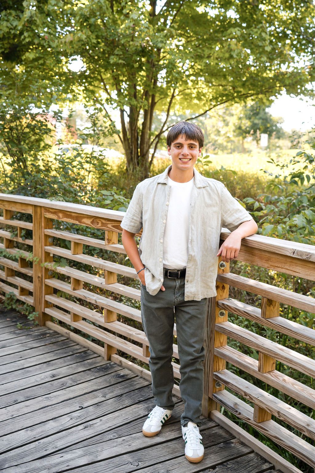 Smiling teen boy in casual outfit standing on wooden bridge in lush green park on sunny day.