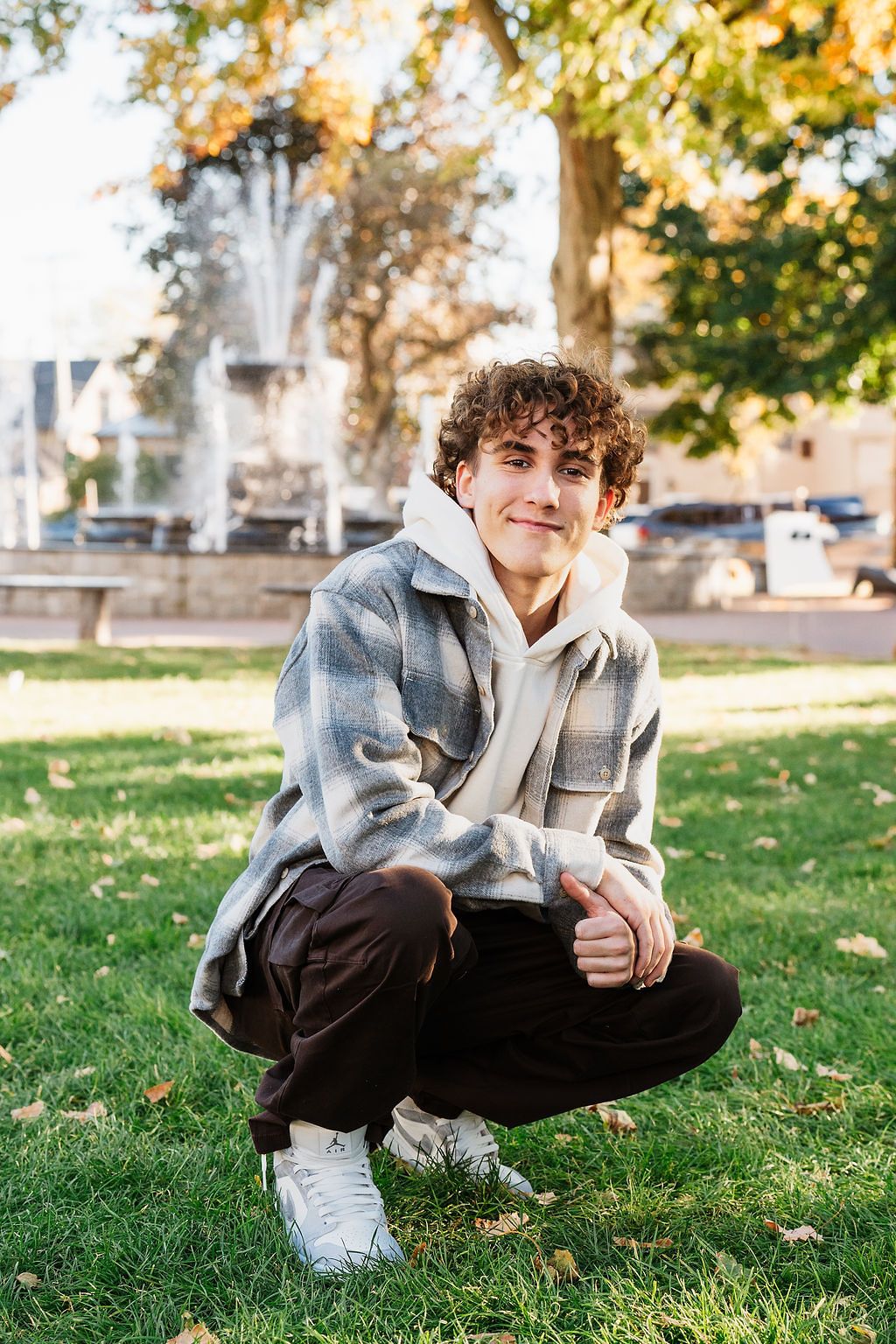 Young man with curly hair crouching on grass in a park, wearing a plaid jacket and white sneakers.