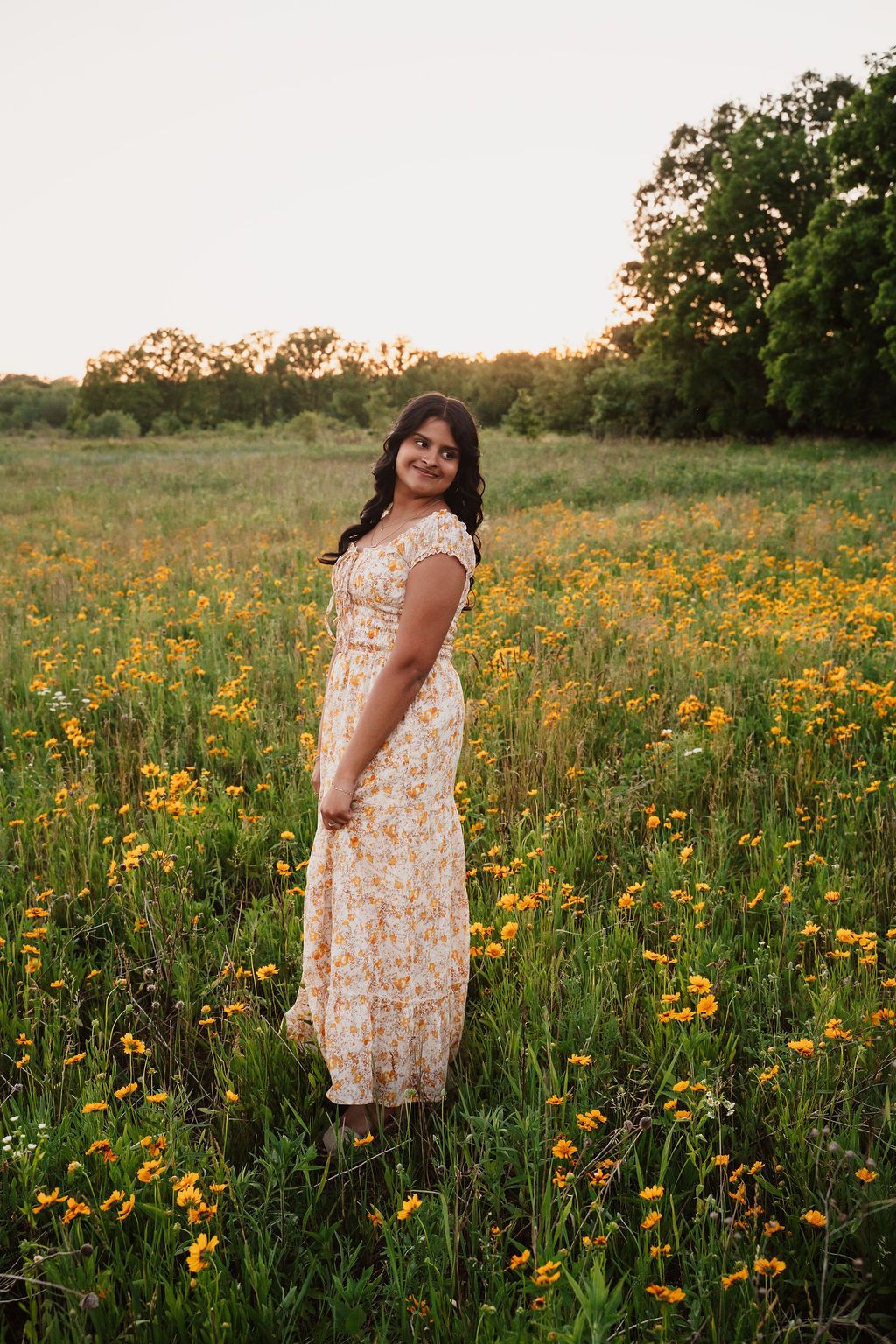 Young woman in floral maxi dress standing in a sunny wildflower meadow with yellow blooms and trees at golden hour.
