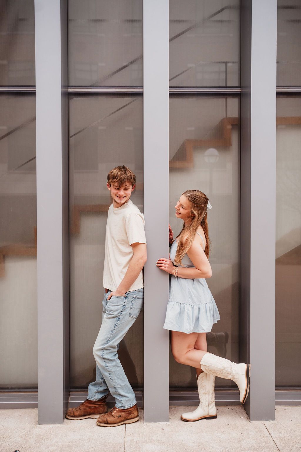 A young couple poses against modern building columns, boy in jeans and cream tee, girl in blue dress and white boots.