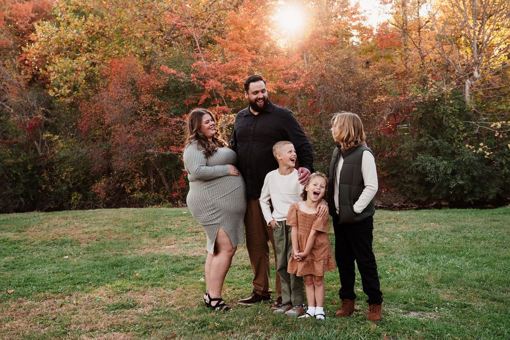 Family of five posing outdoors in fall foliage, parents with three children smiling on green grass with colorful autumn trees.