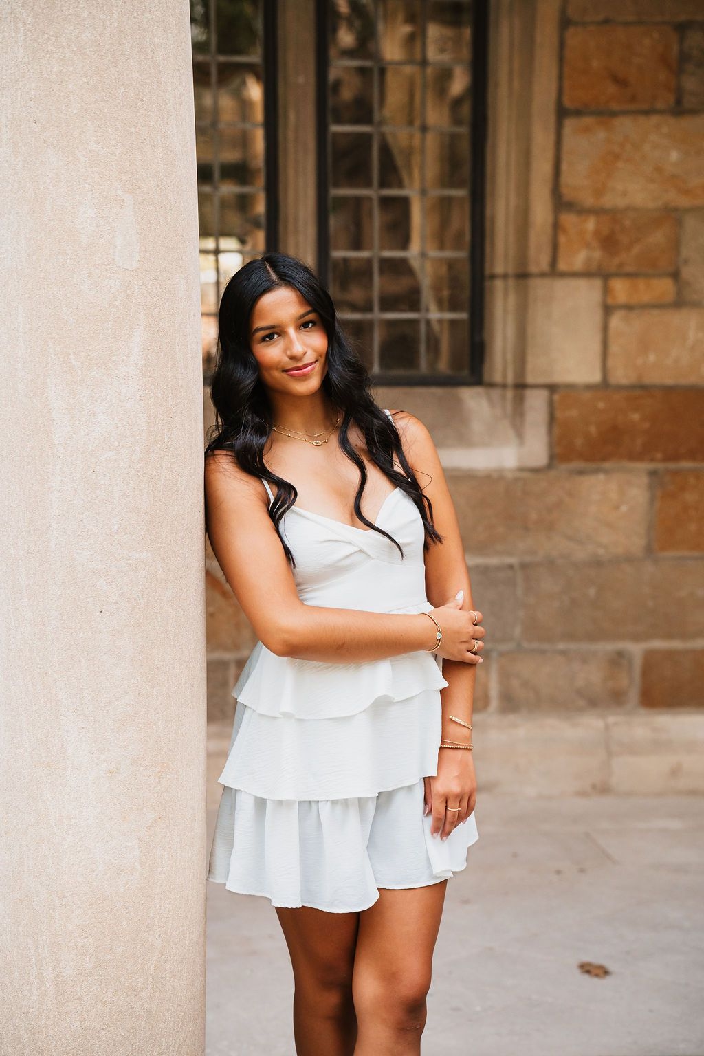 Woman in white ruffled dress poses confidently against stone building with arched windows.