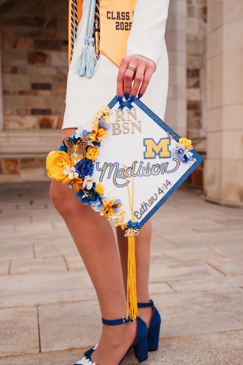 Decorated graduation cap with blue and yellow flowers and tassels displayed against a stone wall background.