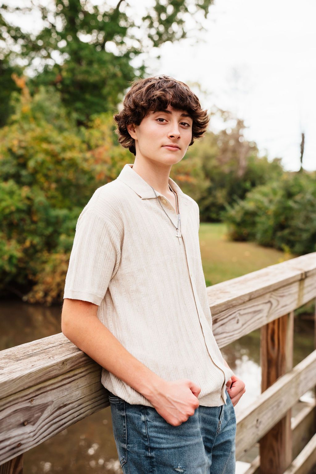 Teen boy in beige shirt and jeans leaning on wooden bridge railing outdoors.