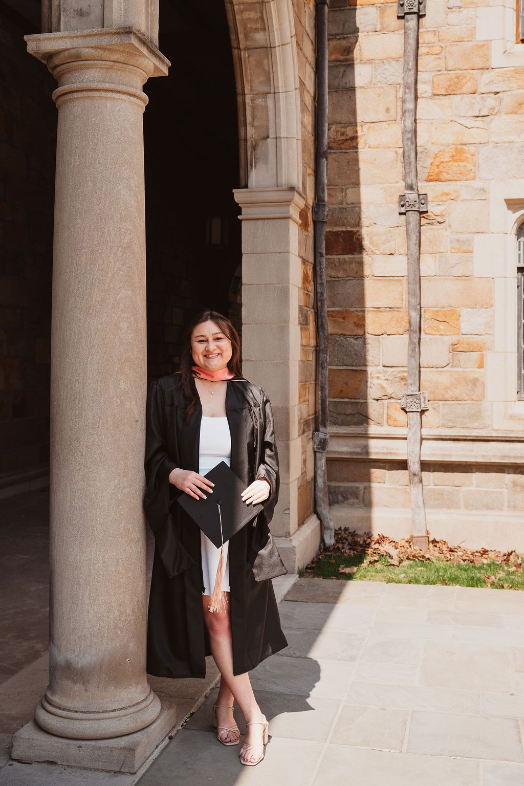 Graduate in black gown with pink hood leans against stone column at University of Michigan.
