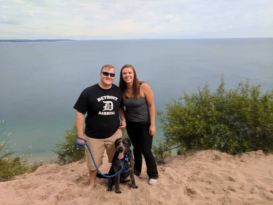 Couple posing with their dog on sandy dunes overlooking Lake Michigan on a cloudy day.