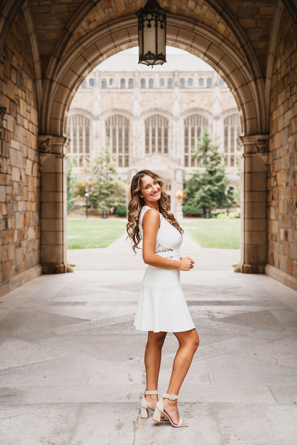 Person in white dress posing under Gothic stone archway at university campus. Ann Arbor law quad PCEP Senior Photos, mini sessions