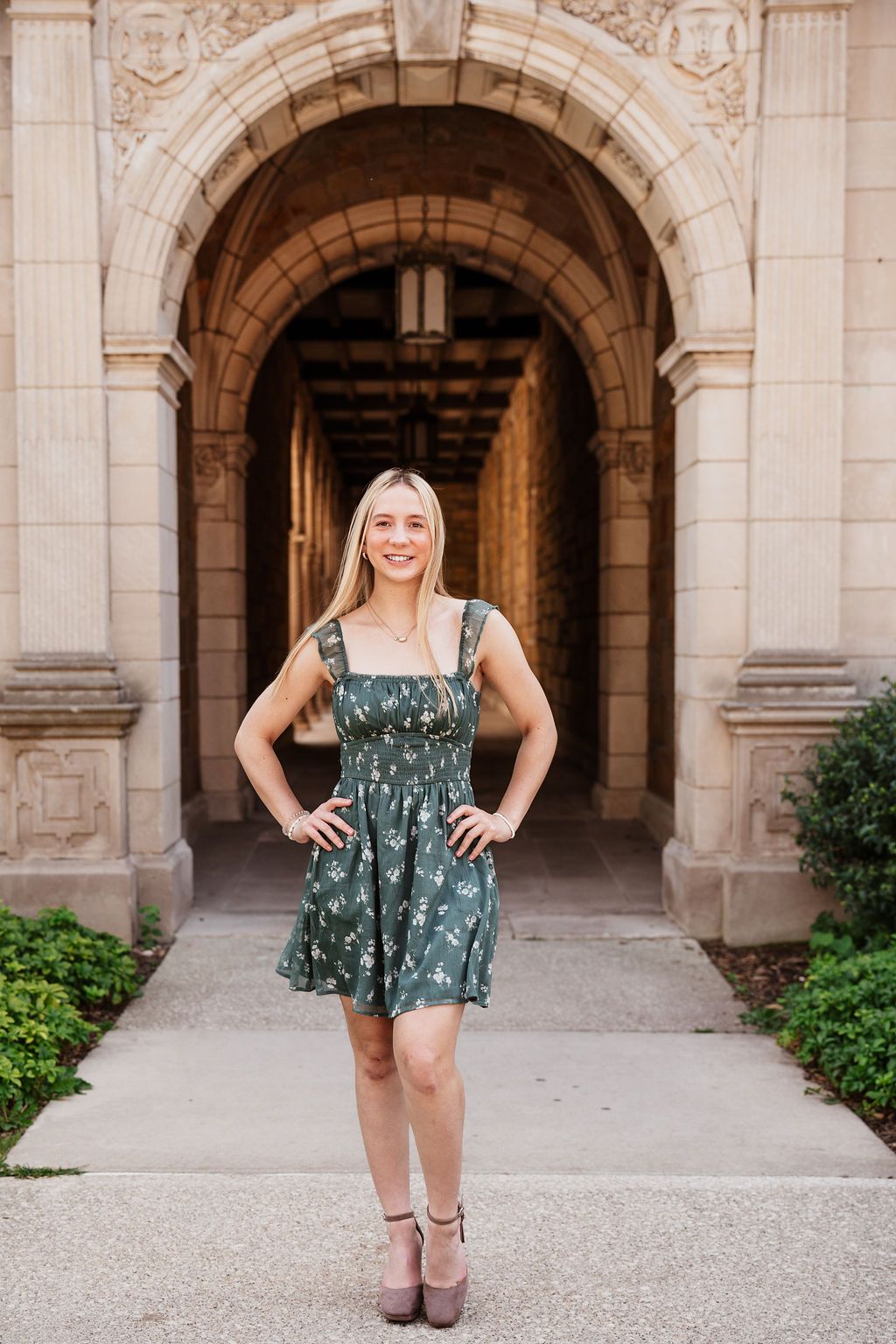 Blonde woman in green floral dress posing under ornate stone archway corridor.