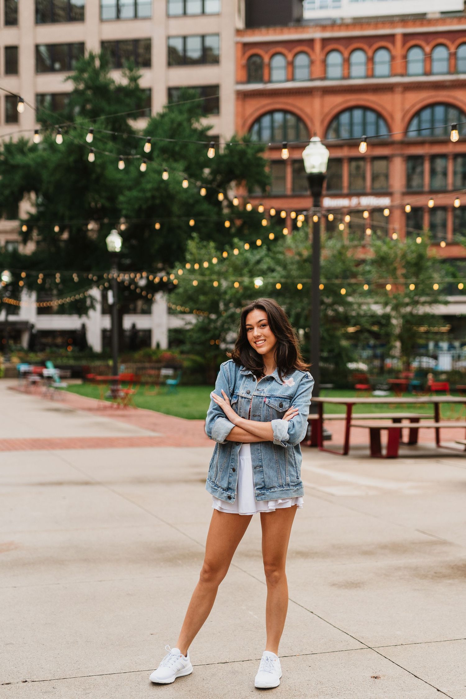 Young woman in denim jacket and white shorts stands confidently in urban plaza with string lights and brick buildings.
