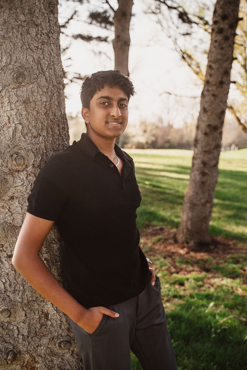 Young man in black polo shirt leaning against tree in park for outdoor senior portrait.