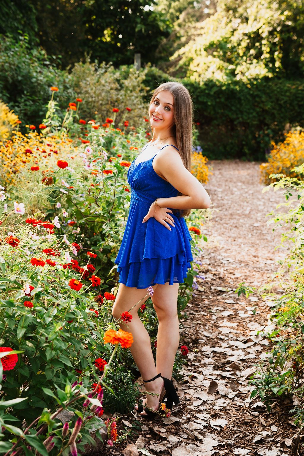 Young woman in blue dress posing in colorful garden with red flowers and green trees.