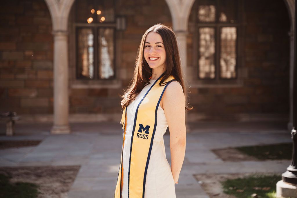 Smiling young woman in University of Michigan graduation stole posing outdoors near brick building.