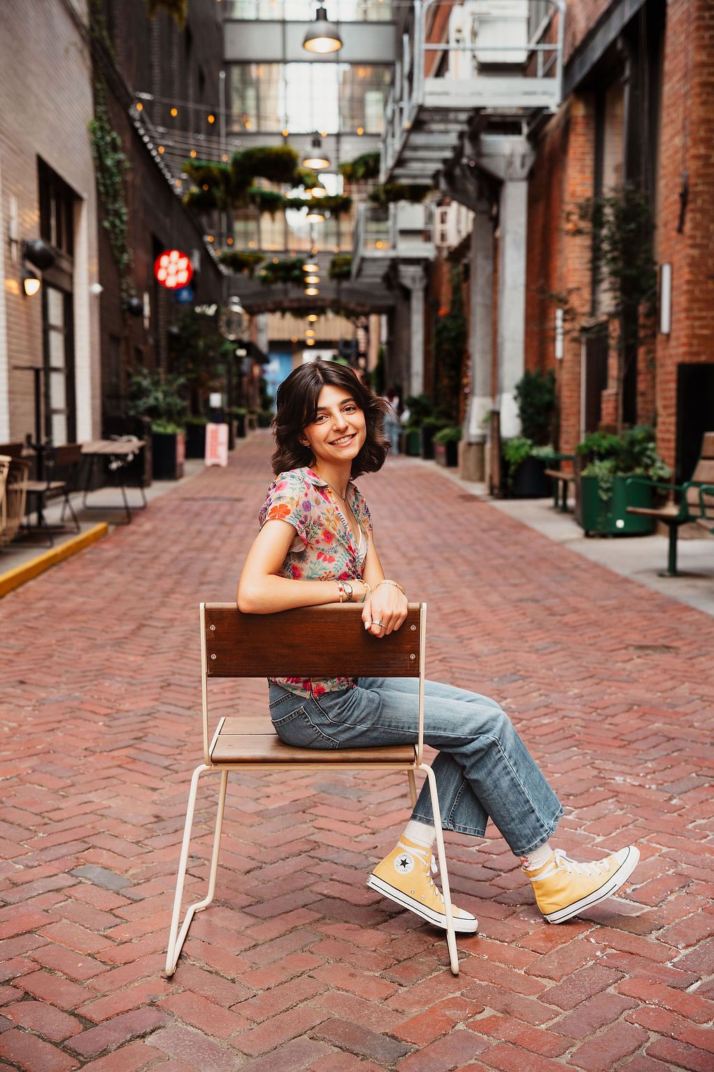 Young woman sitting backwards on a chair in a brick alley, wearing jeans, floral top, and yellow Converse sneakers.