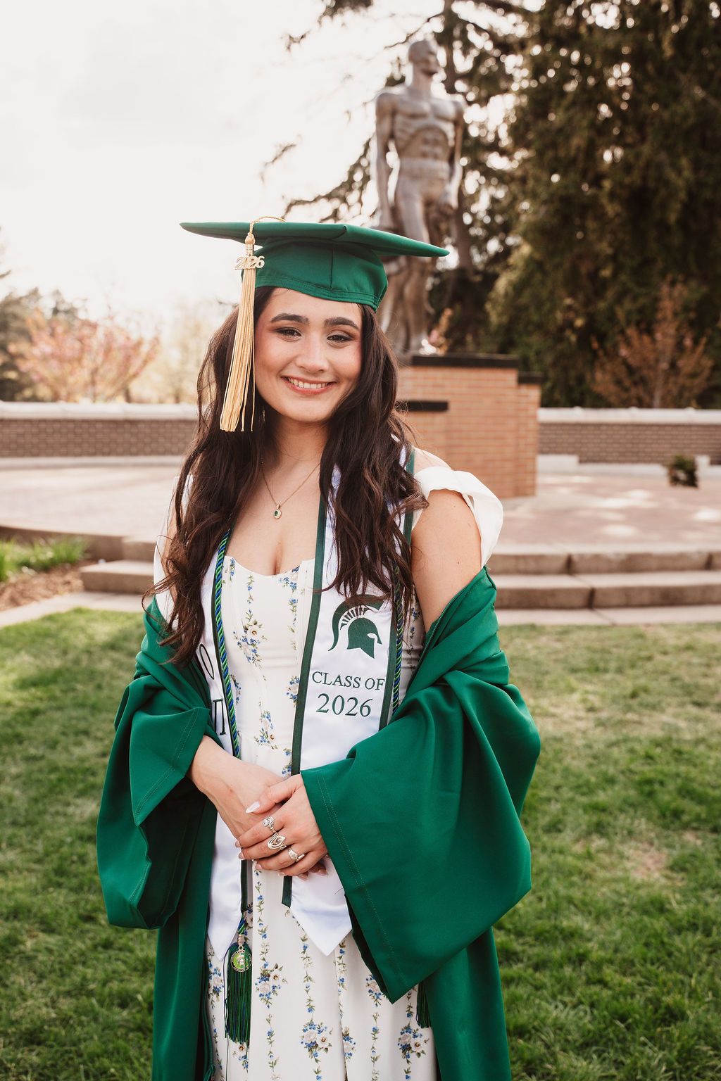 Smiling woman in green Michigan State Class of 2026 graduation cap and gown outdoors near campus statue.