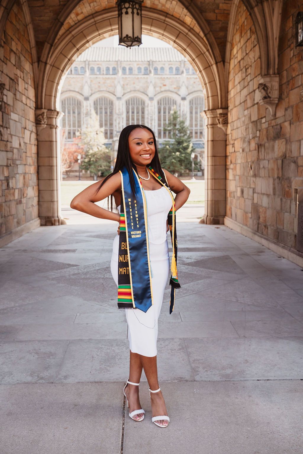 Smiling graduate in white dress wearing colorful graduation stoles poses under stone archway at university campus.