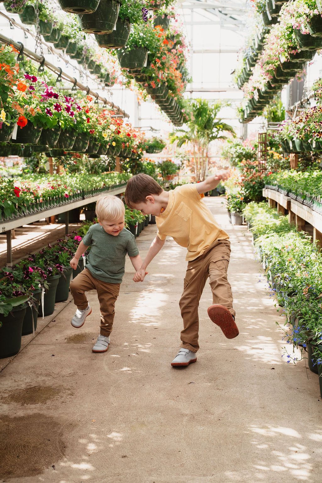 Two young boys joyfully playing and holding hands in a sunlit greenhouse filled with colorful flowers.