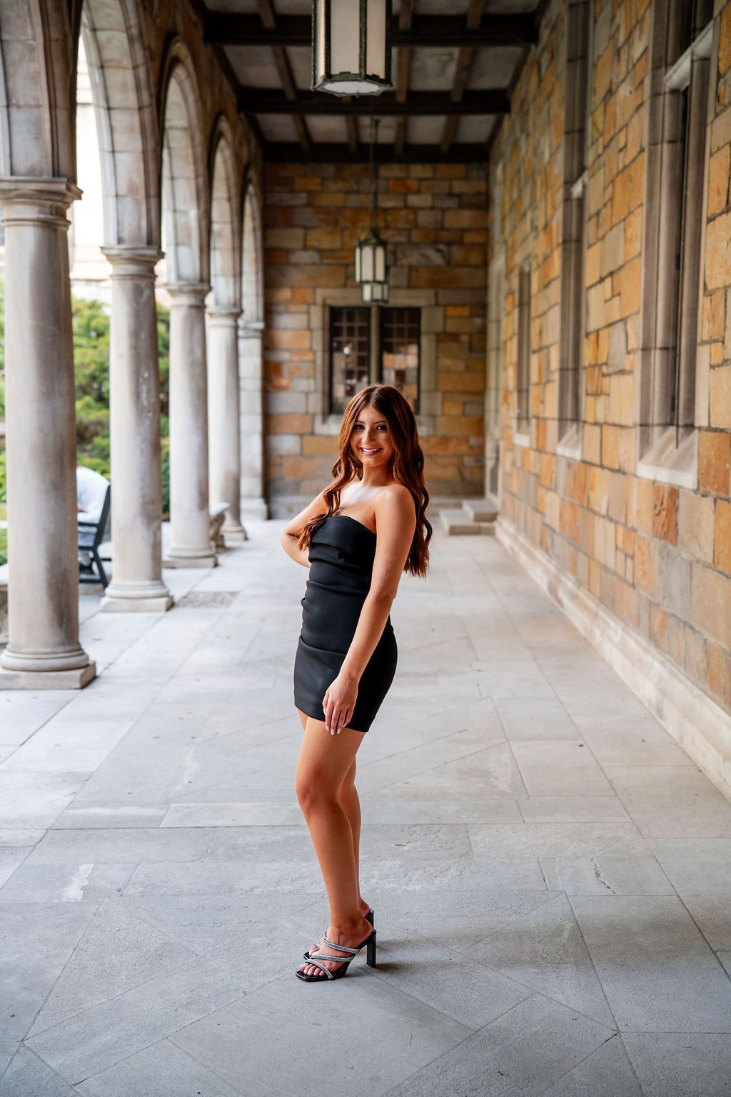 A figure poses in a black outfit against a stone colonnade with arched walkways and brick walls.