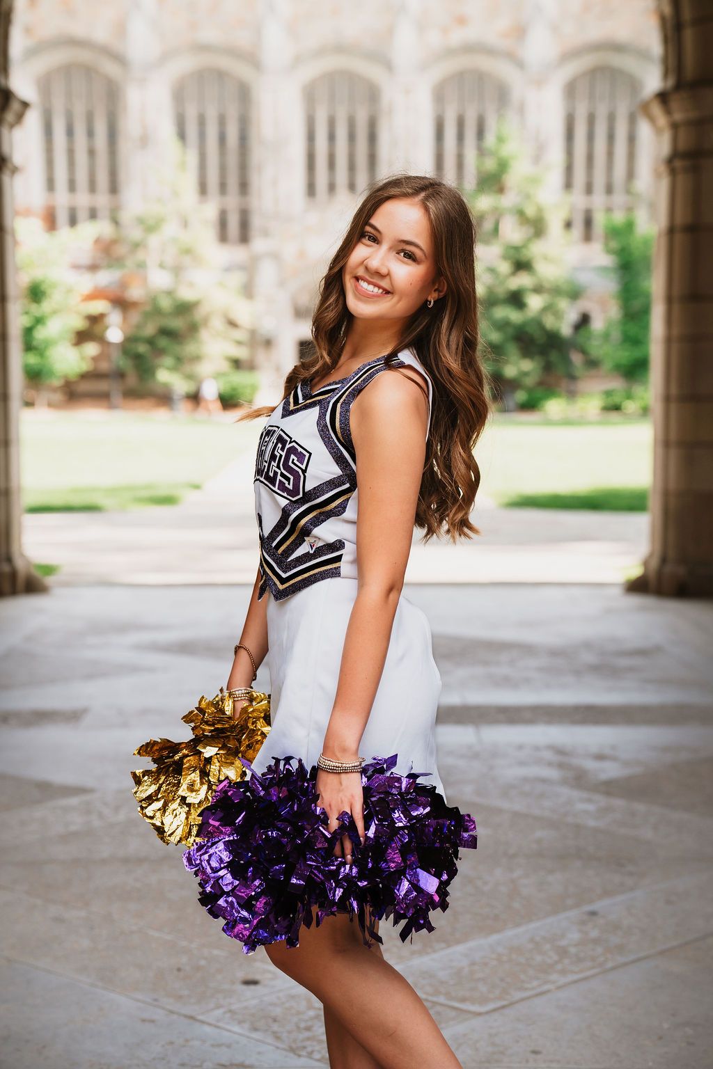 Smiling cheerleader in white uniform holding gold and purple pom-poms poses in an arched stone courtyard.