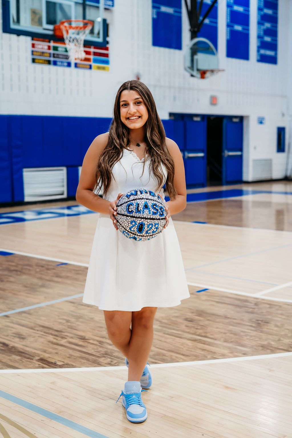 A person in a white dress and blue sneakers poses on a basketball court with blue walls and wooden flooring.