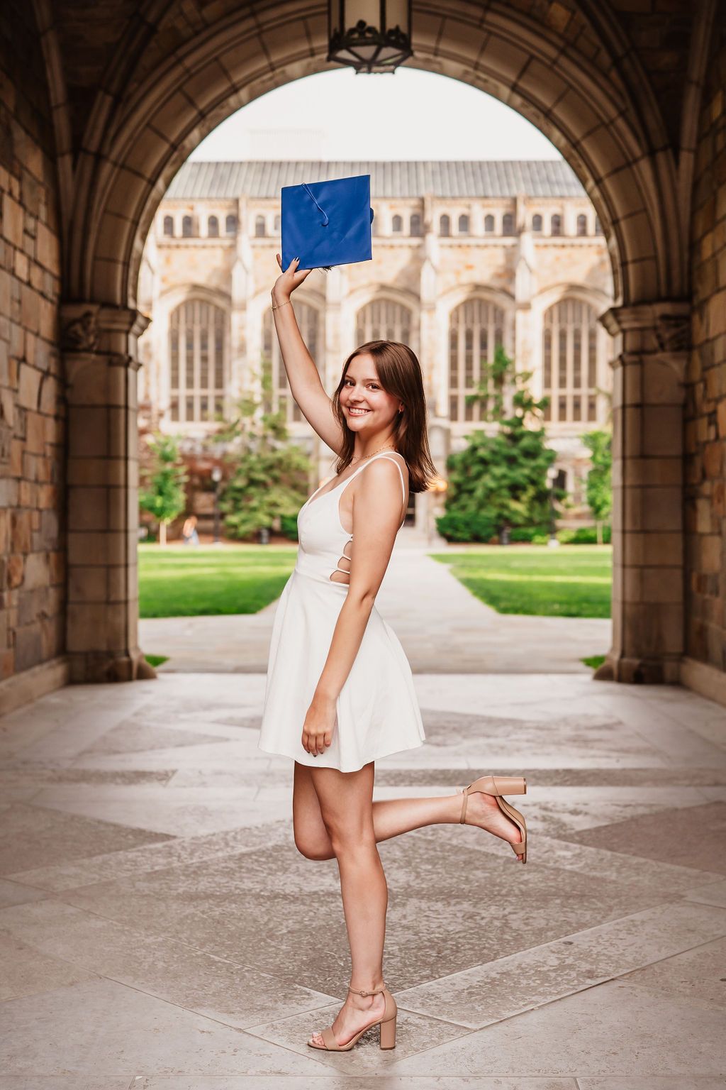 Graduate in white dress strikes elegant pose under ornate stone archway with university courtyard behind.