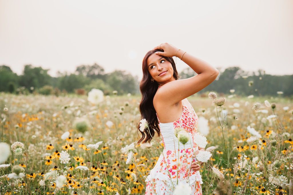 A woman in a floral summer dress poses joyfully in a wildflower meadow on a sunny day.